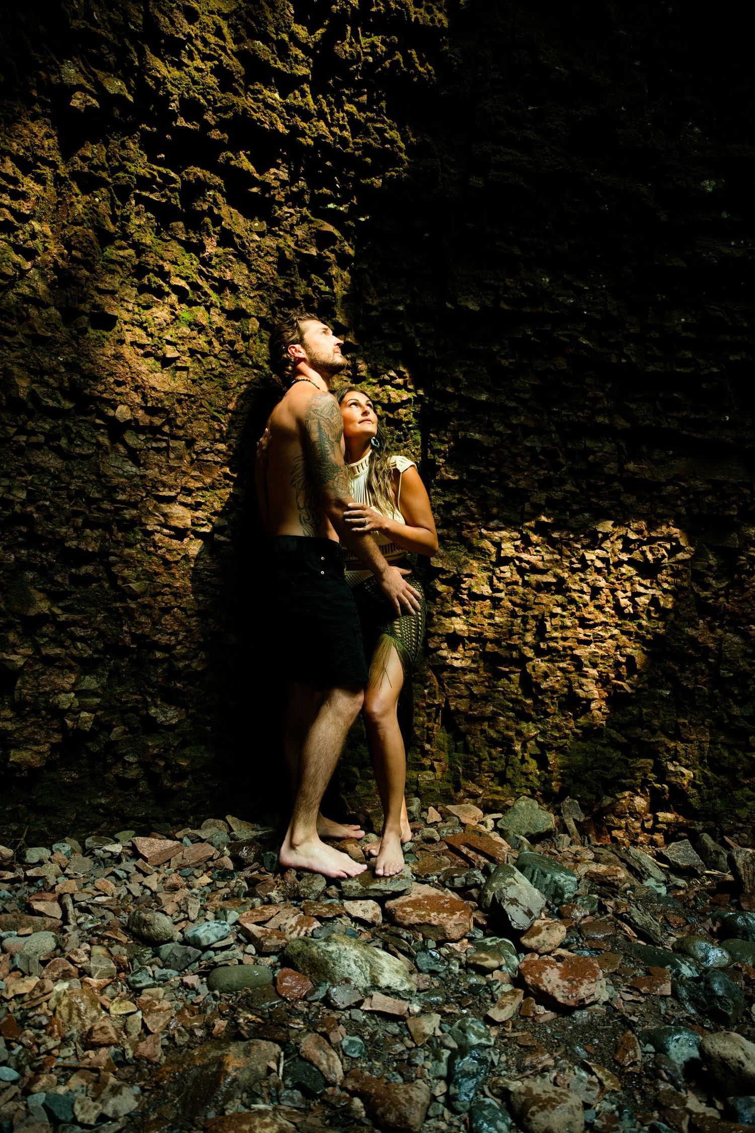 Couple embraces against mossy rock wall in dark cave standing on scattered stones.