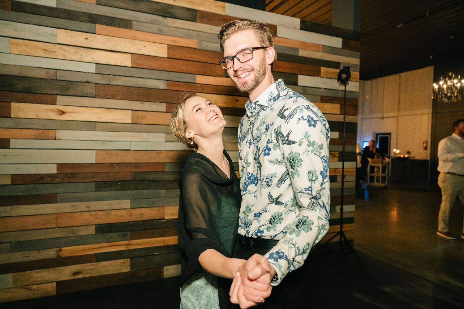 Couple smiles and holds hands while dancing in front of a reclaimed wood wall at a reception.