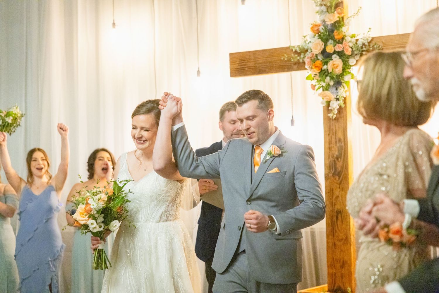 Joyful bride and groom walk back down aisle after wedding ceremony beside rustic wooden cross.