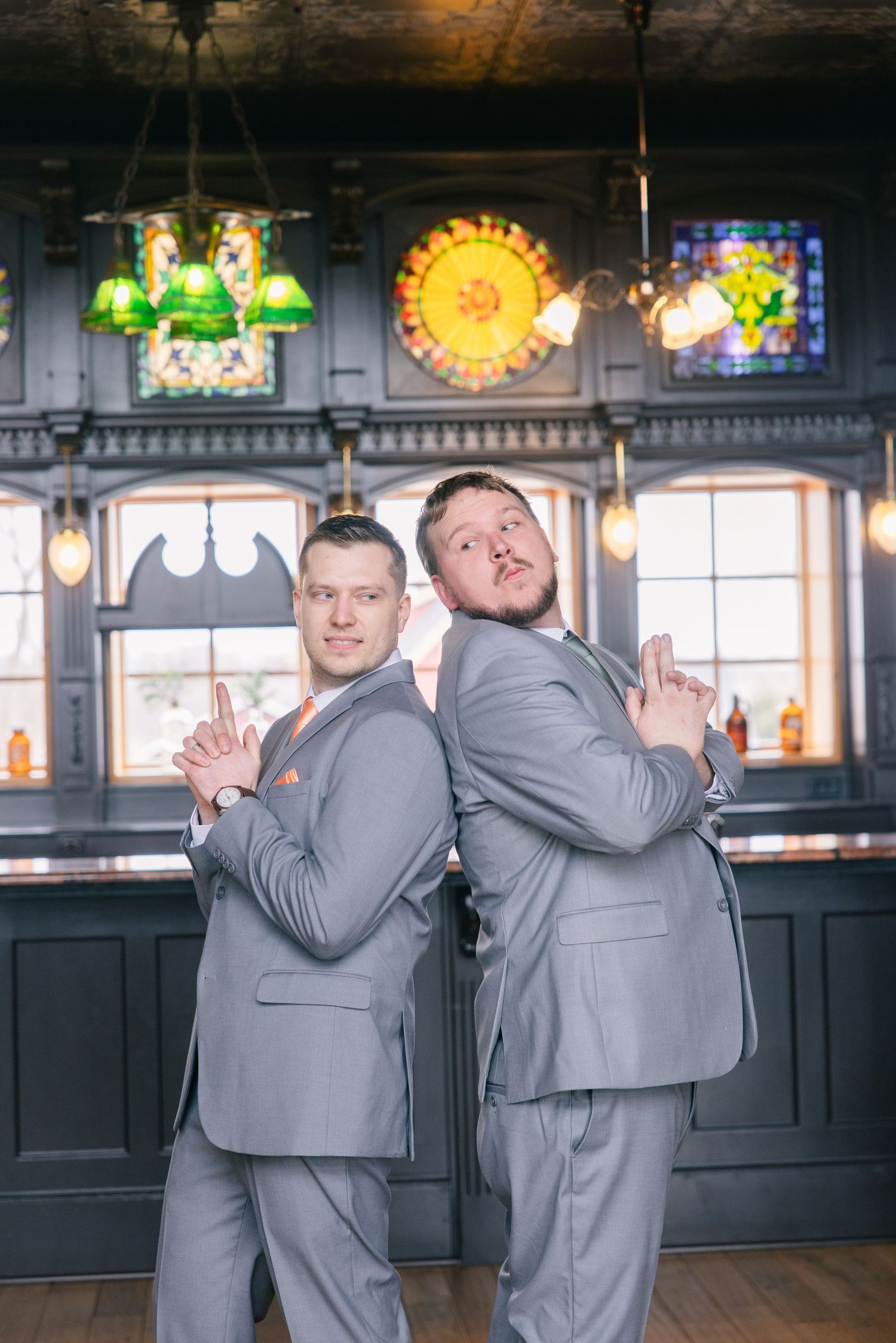Two grooms in gray suits strike a fun pose back-to-back in a vintage bar with stained glass decor.
