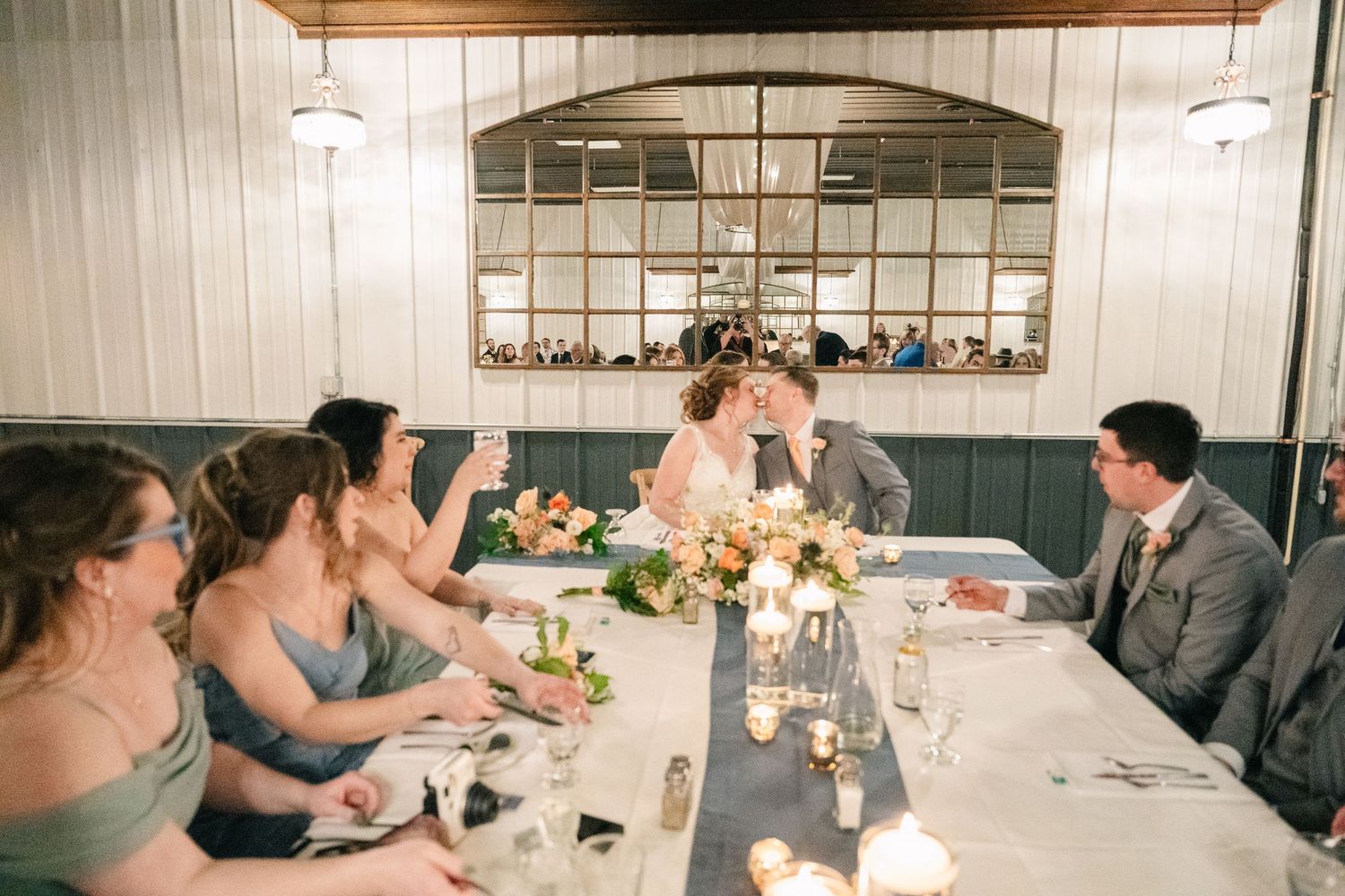 Bride and groom share kiss at sweetheart table surrounded by bridesmaids and floral centerpieces.