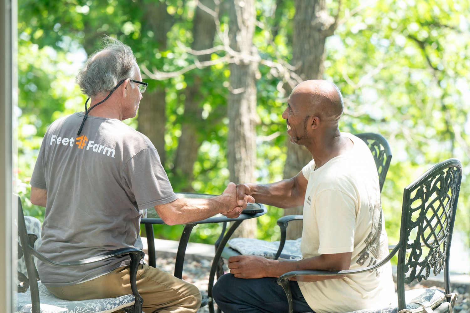 Two men having a conversation outdoors on a porch, one wearing a Feed the Farm shirt, surrounded by lush green trees.