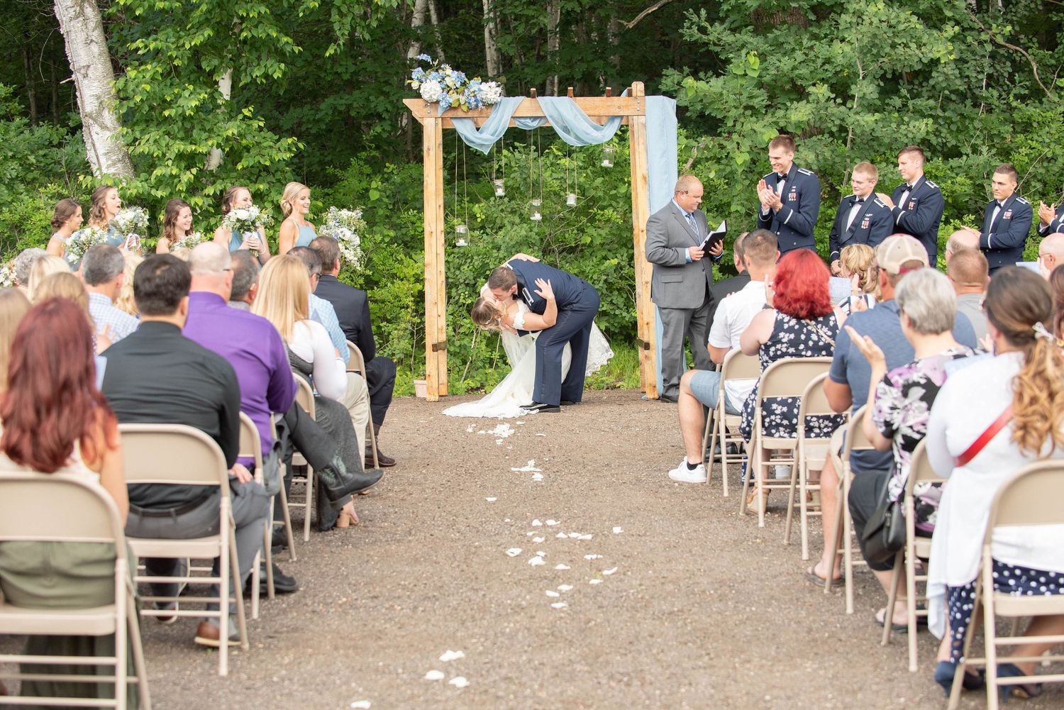 Wide view of outdoor wedding ceremony with guests seated in white chairs surrounded by trees. Zimmerman MN Weddings