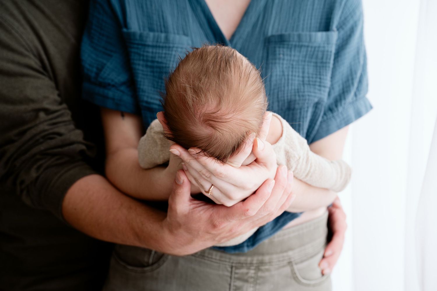 Minnesota Best newborn photographer -A parent in blue shirt cradles a tiny newborn baby wrapped in light pink blanket against window light.