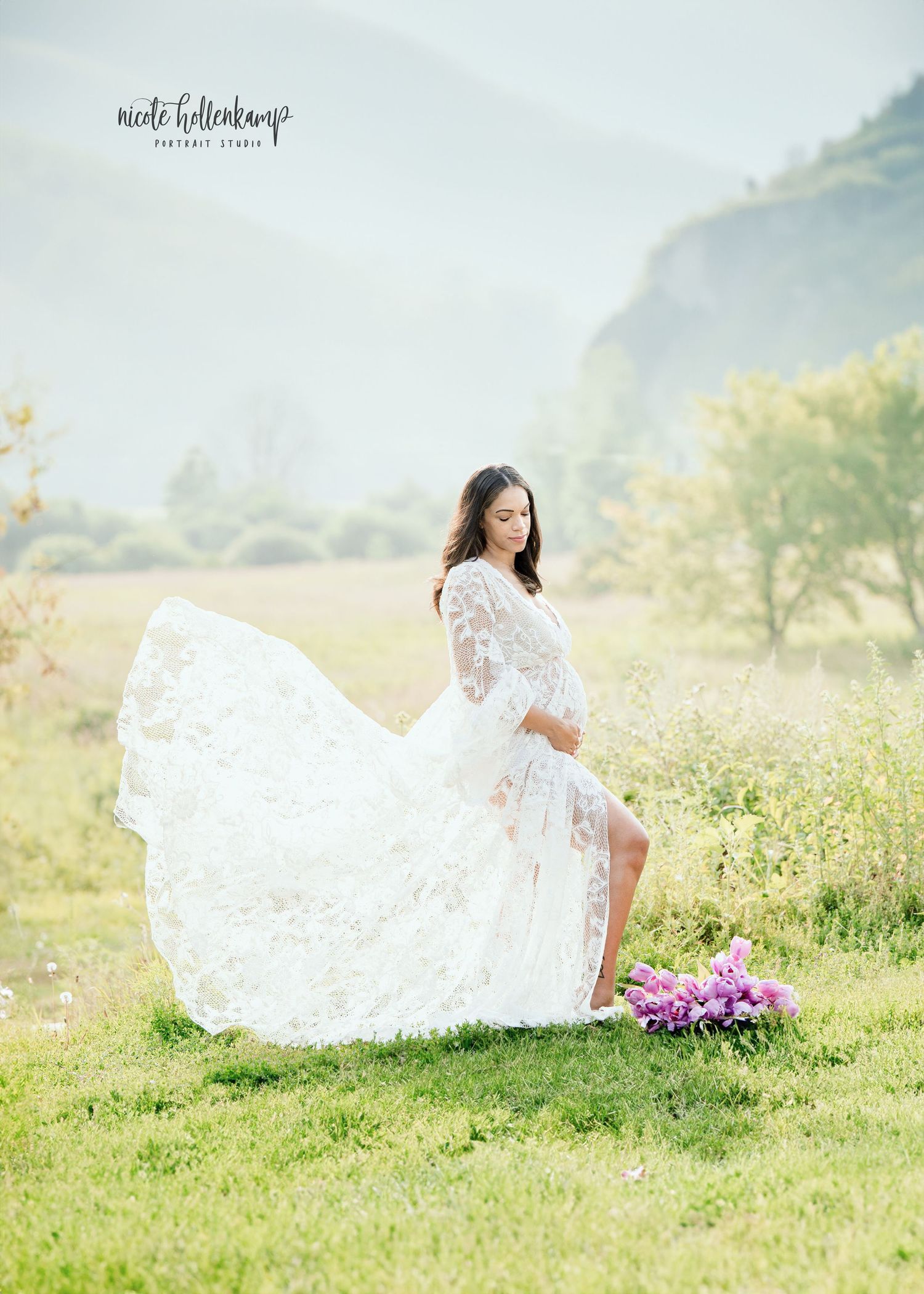 Pregnant woman in flowing white lace gown poses in sunlit meadow with mountain backdrop and pink flowers nearby.