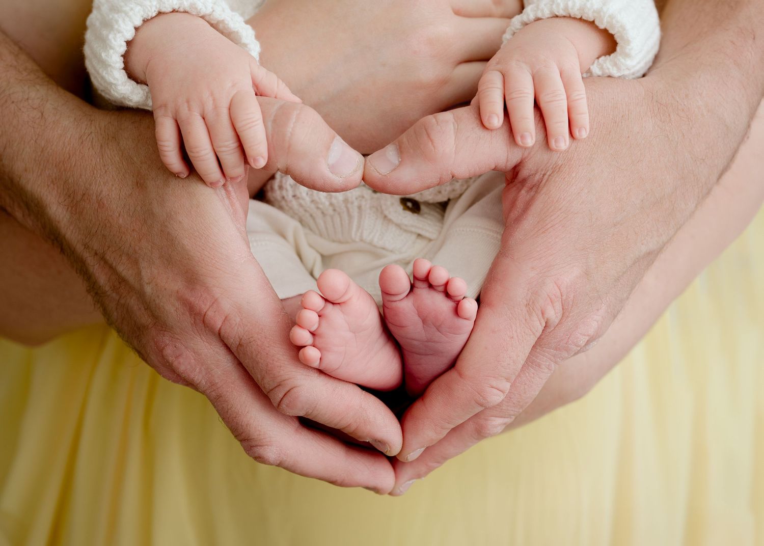Adult hands gently hold tiny newborn baby feet against a soft yellow blanket.