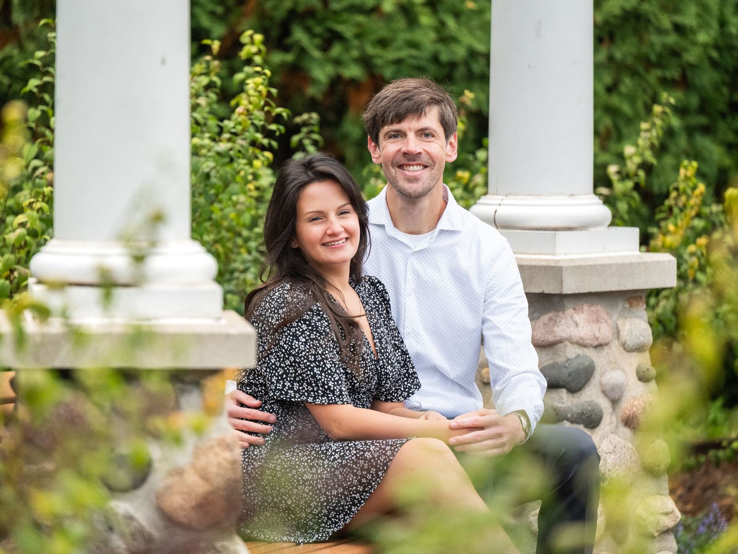 In central Minnesota a Couple posing together outdoors against white pillars with lush greenery in the background.