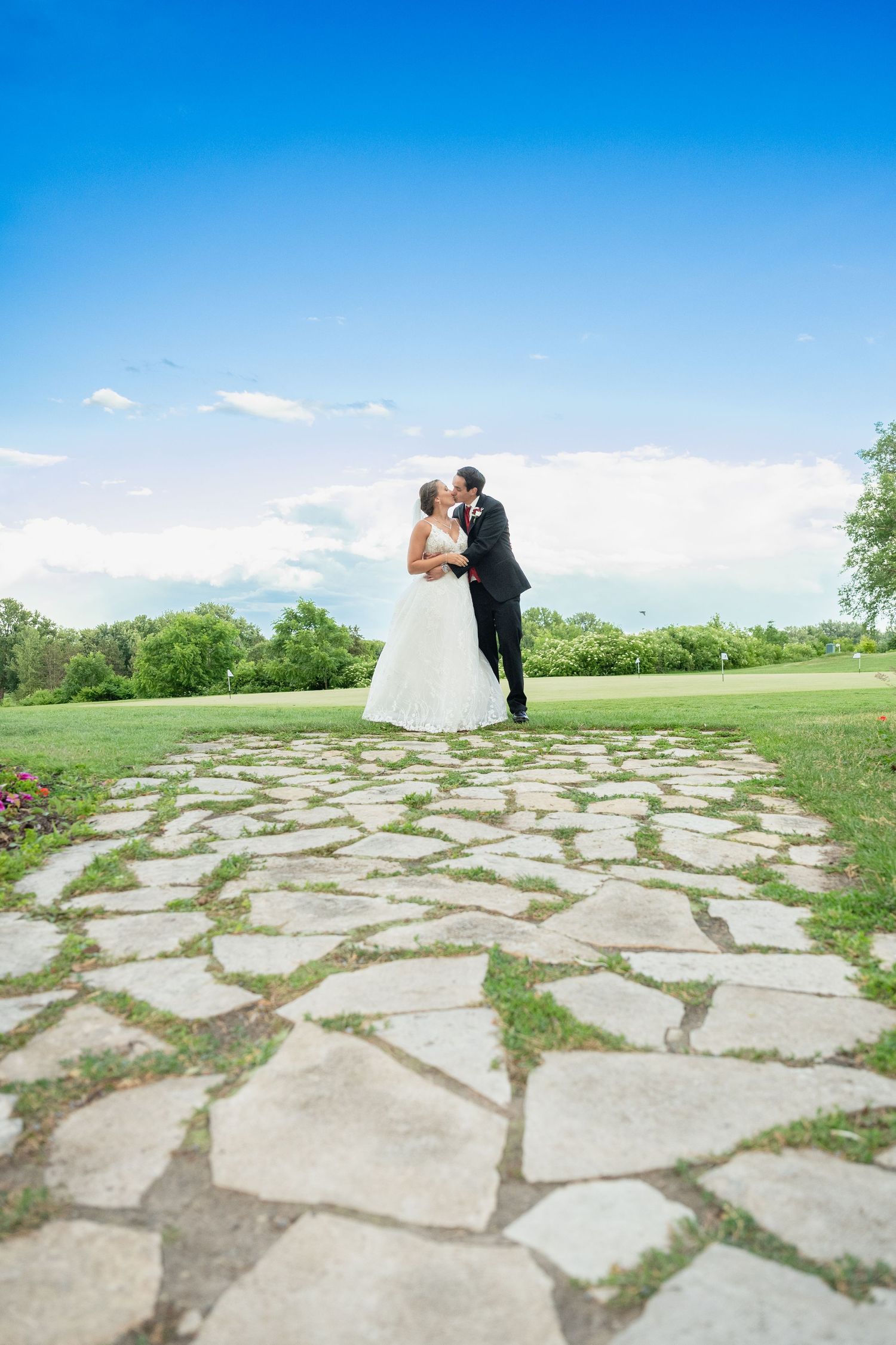 Bride and groom share a kiss on a stone pathway surrounded by lush green grass under a bright blue sky.