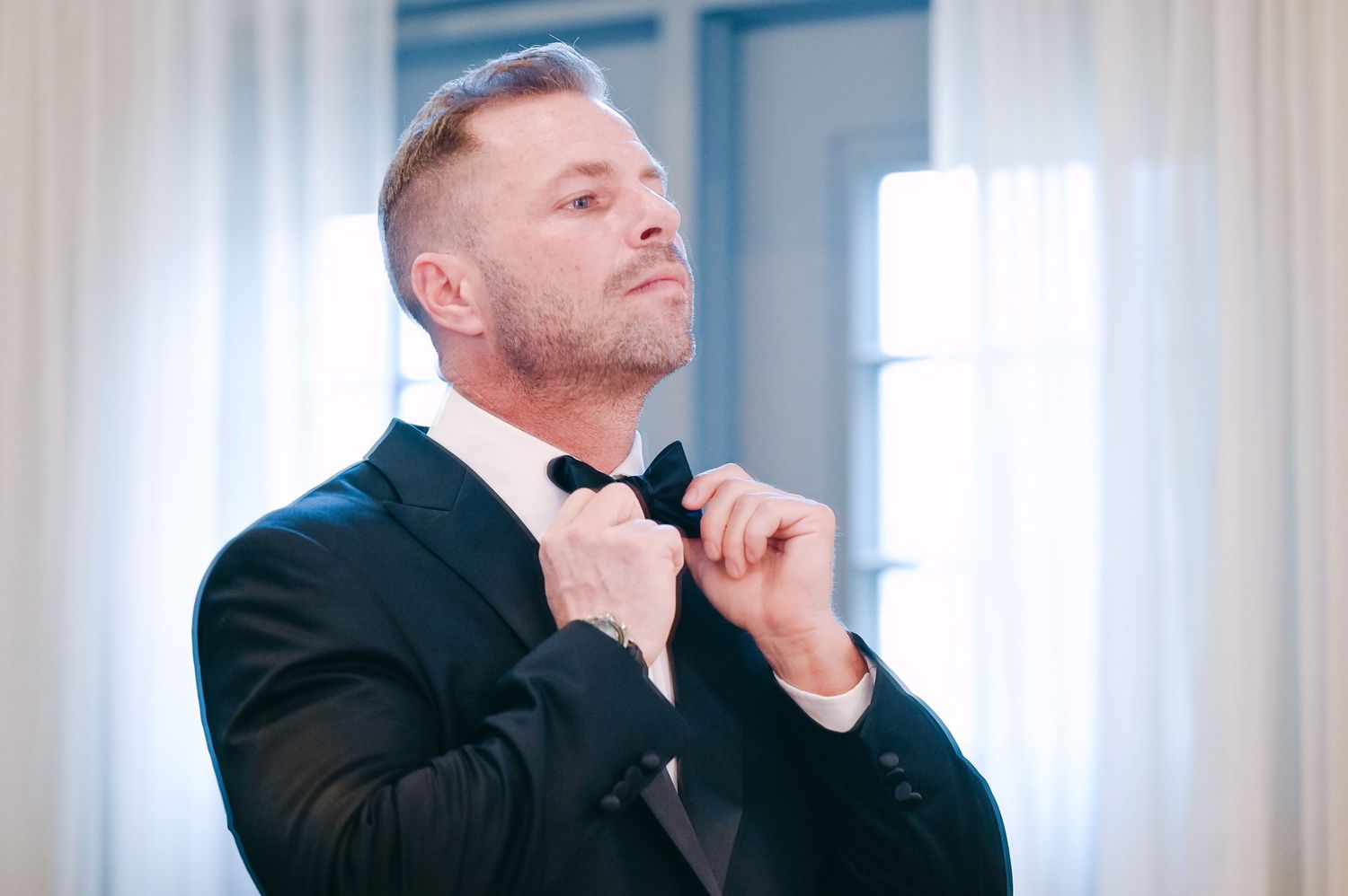A person in formal attire touches their collar while standing in a bright room with exposed brick walls.