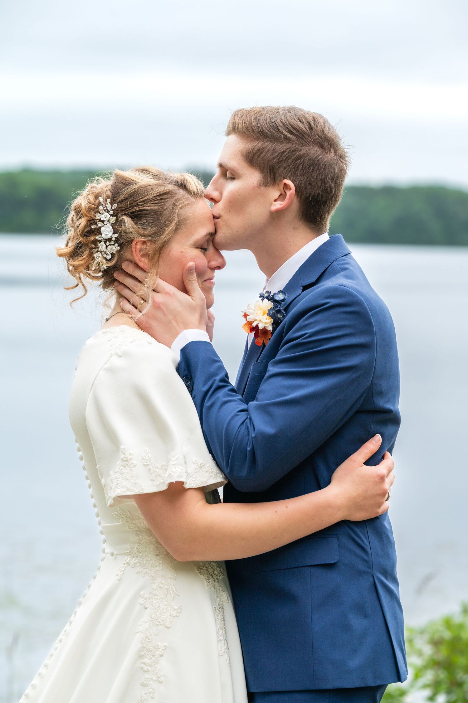 A couple shares a romantic moment in wedding attire by a lakeside, with the bride in white and groom in navy blue.
