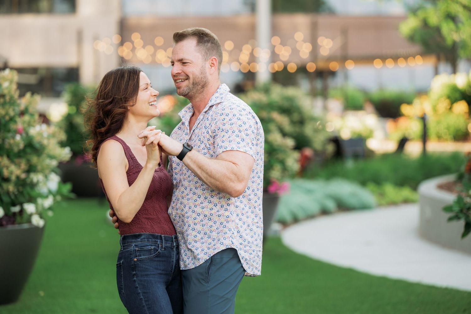A couple dances and laughs together in a garden with string lights and greenery in the background.