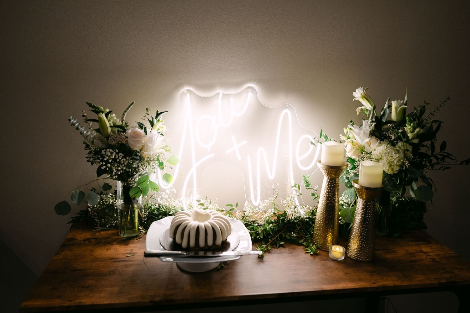 Wedding dessert display featuring a bundt cake, neon light sign, and greenery on wooden table.