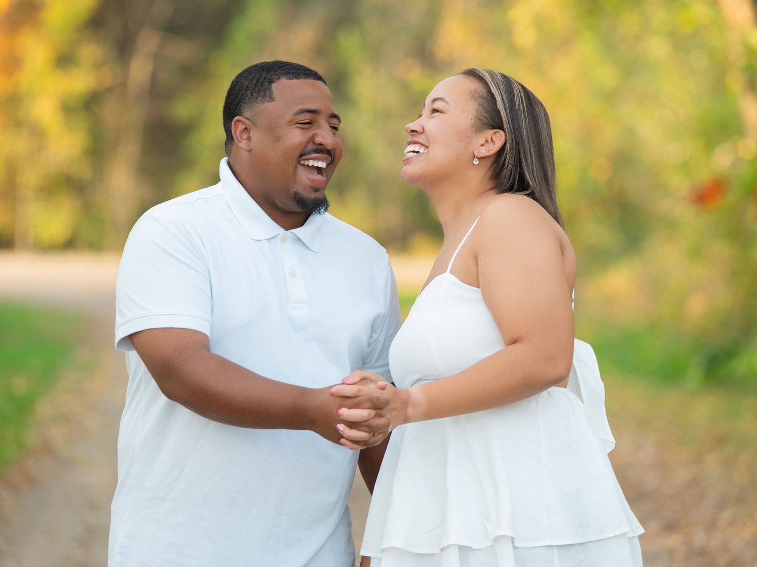 A joyful couple wearing white outfits laughs together while holding hands outdoors during a sunny day.