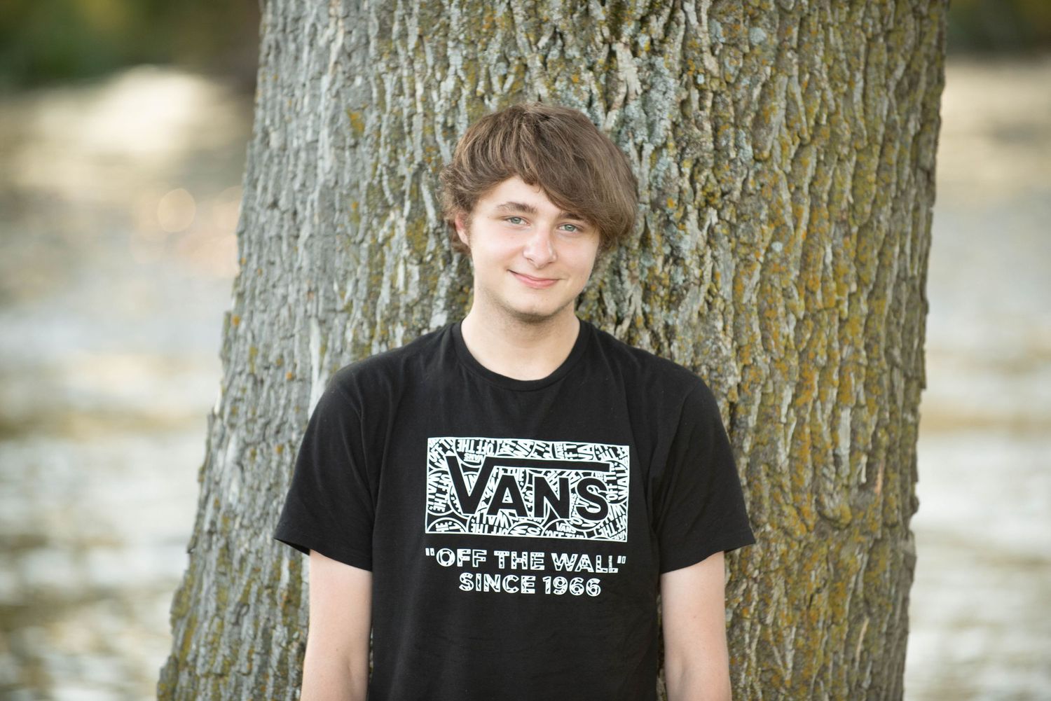 Person wearing black Vans t-shirt leans against tree trunk in outdoor portrait session.