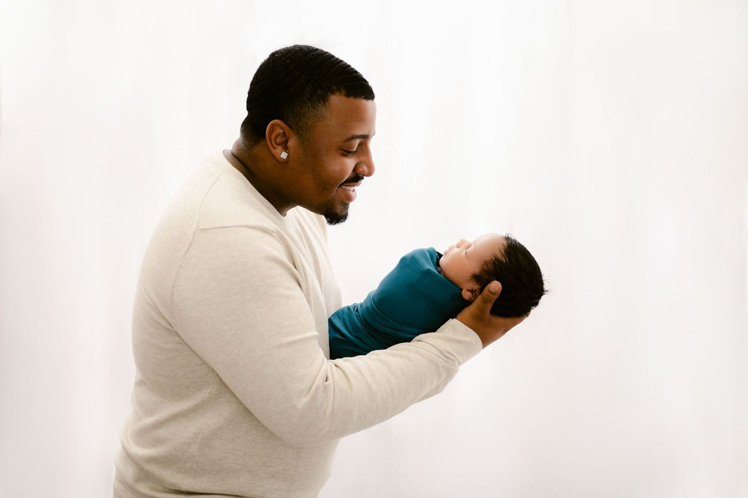 St Cloud newborn Photos of Proud father in cream sweater smiling at his newborn baby wrapped in a teal swaddle against white background.