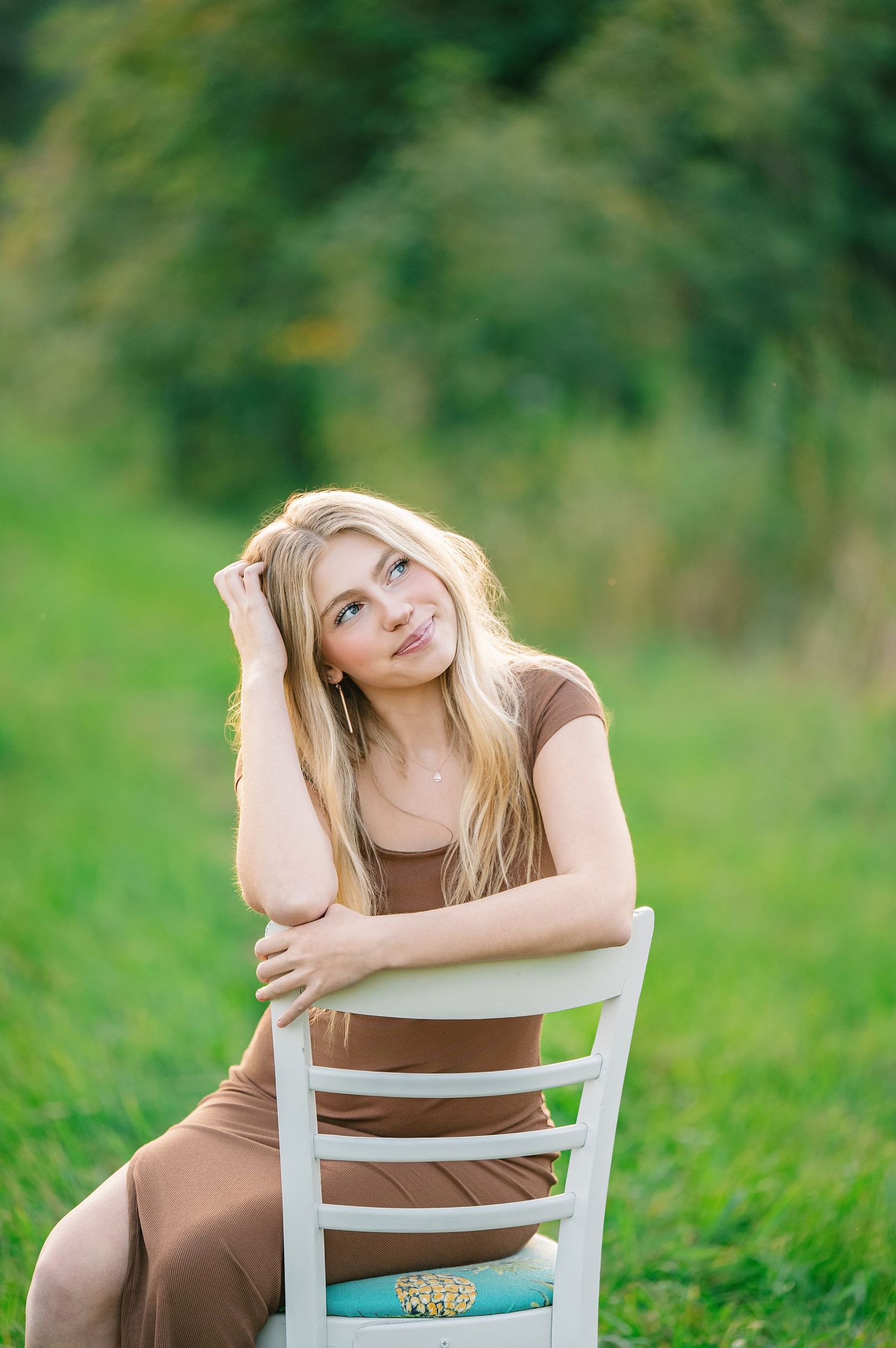 Central MN Senior Photography - Young blonde woman in brown dress sitting on white chair outdoors in green field, looking up thoughtfully.