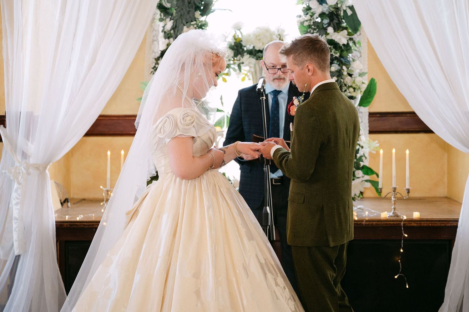 Central MN Wedding Photos | Bride placing wedding ring on groom's finger during ceremony with officiant and white floral arch.