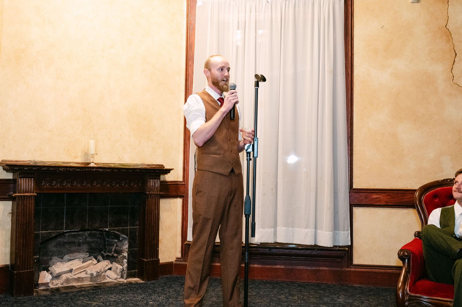 Central MN Wedding Photos | Man in brown vest speaks at microphone near fireplace at wedding reception.
