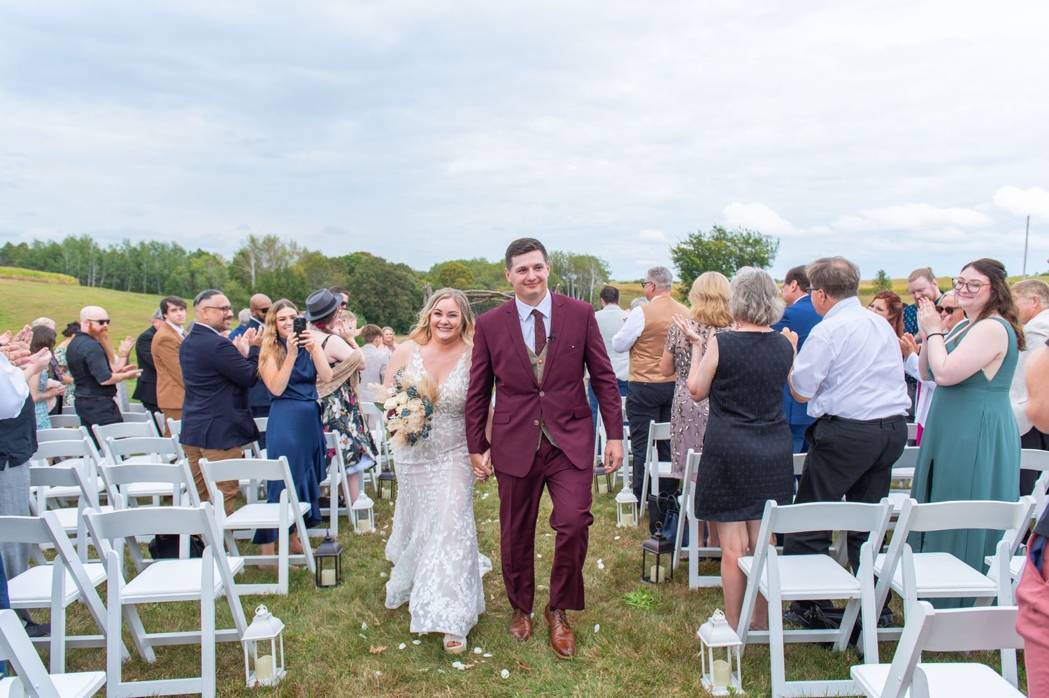 Central MN Wedding Photography - Newlyweds walk down outdoor aisle in burgundy suit and lace wedding dress, guests celebrating around them.