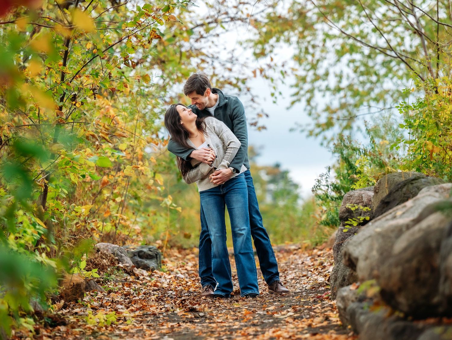 Central MN Engagement Session.  Couple sharing a romantic kiss on a leaf-covered autumn trail surrounded by colorful fall foliage.