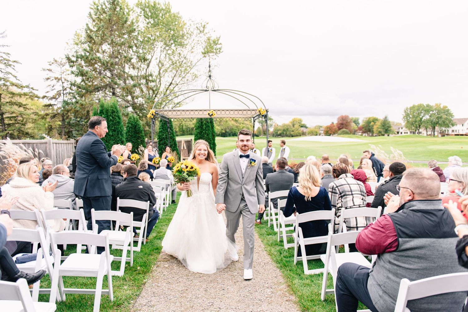 Central MN Wedding Photos - Newlyweds walk back down the aisle together after outdoor wedding ceremony as guests celebrate.