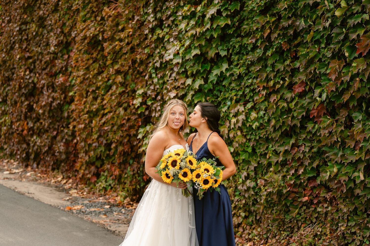Central MN Wedding Photos | Bride and bridesmaid in navy dress smile together holding sunflower bouquets by ivy wall.
