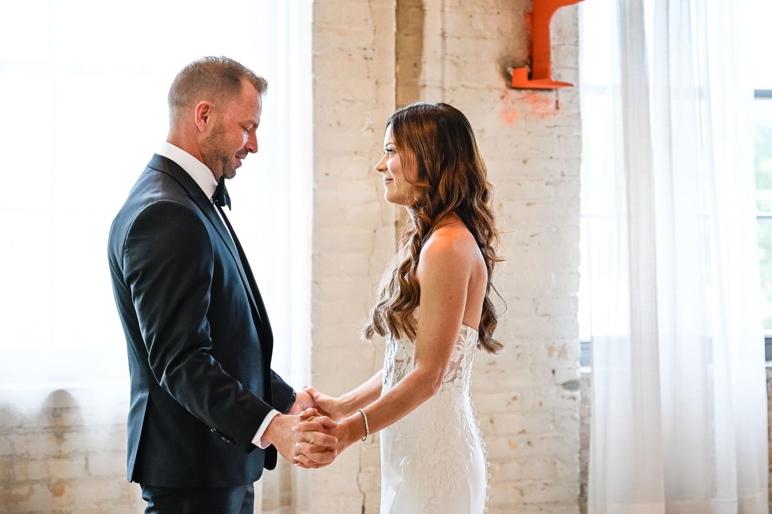 Central MN Wedding Photos | Bride and groom share a tender first look moment, holding hands and smiling in a bright industrial venue.