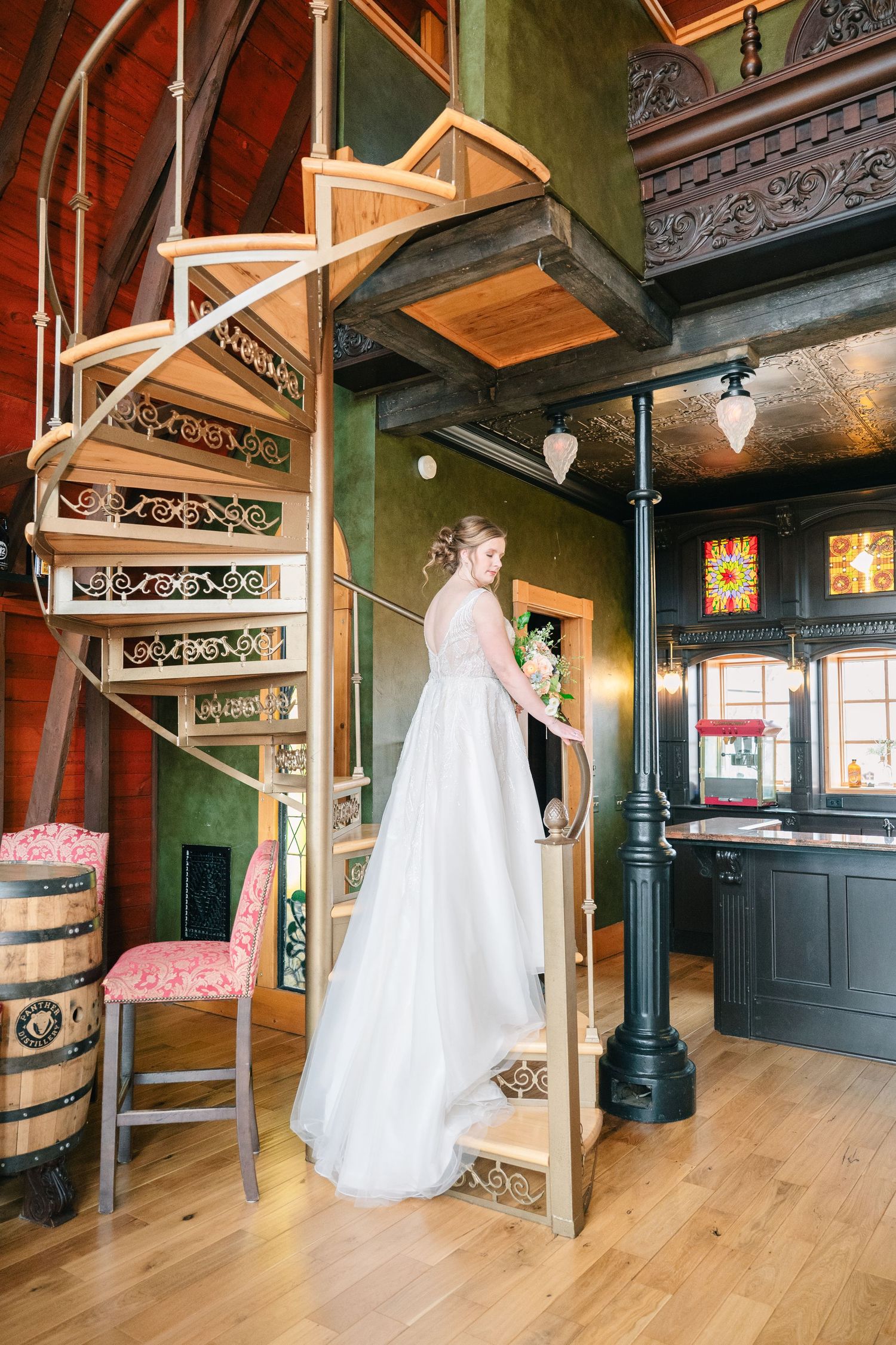 Bride in white gown stands on spiral staircase in ornate venue with green walls and stained glass windows.