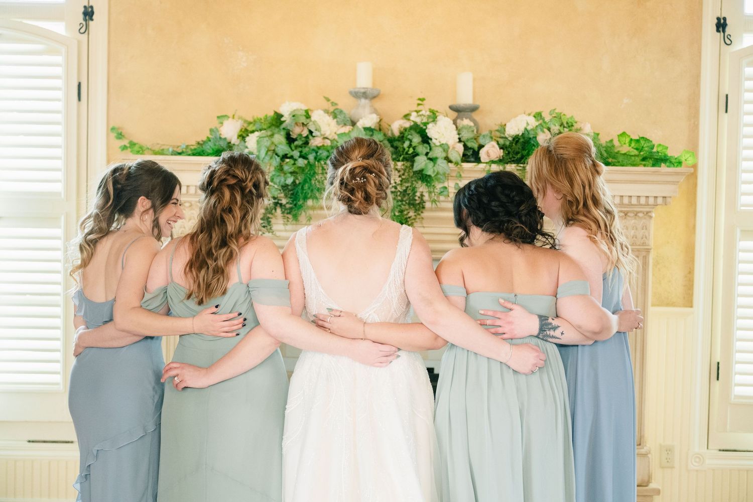 Bride and four bridesmaids in sage green dresses embracing with arms around each other.