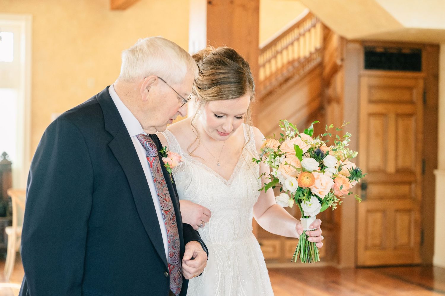 Bride in white gown holding peach bouquet shares tender moment with elderly father indoors.