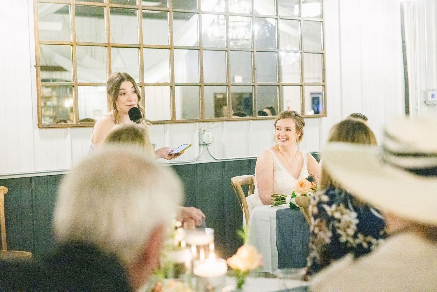 Bridesmaid delivers a heartfelt speech at a wedding reception while the bride laughs joyfully.