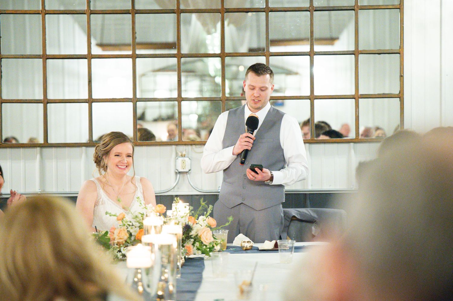 Groom speaks into a microphone at the wedding reception head table beside his smiling bride.