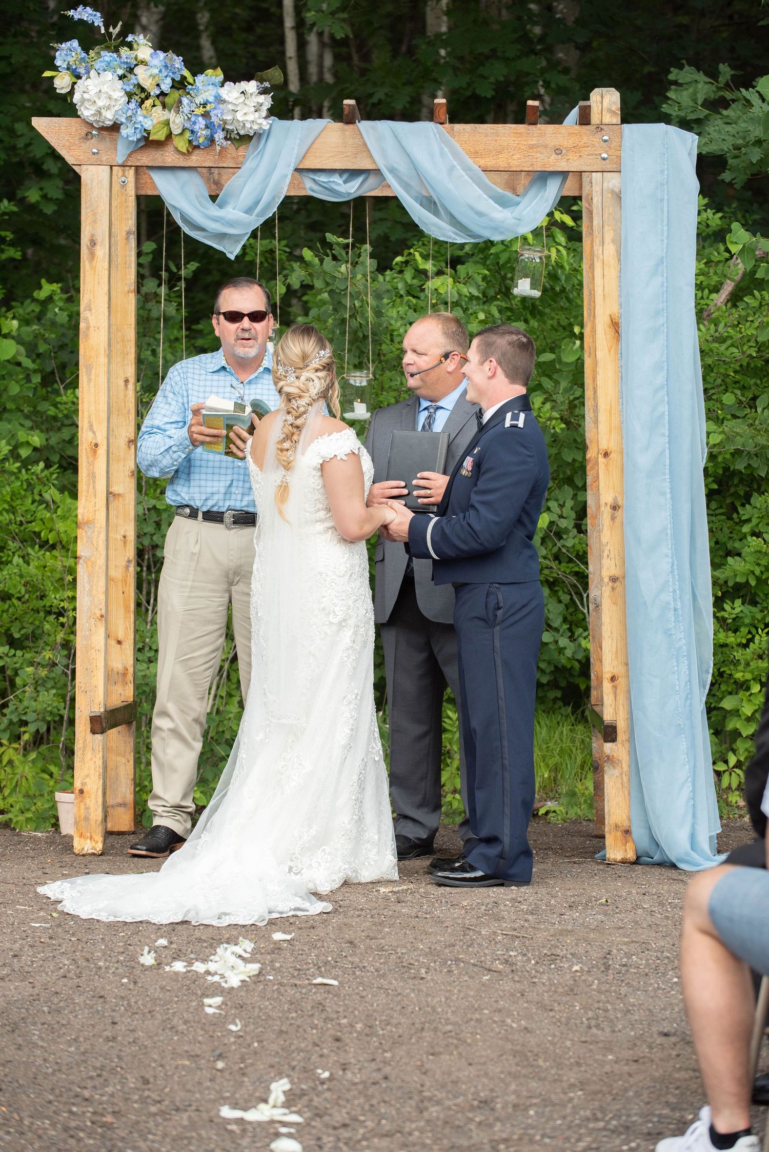 An outdoor wedding ceremony under a wooden arch decorated with light blue fabric and white floral arrangements. Zimmerman MN Weddings