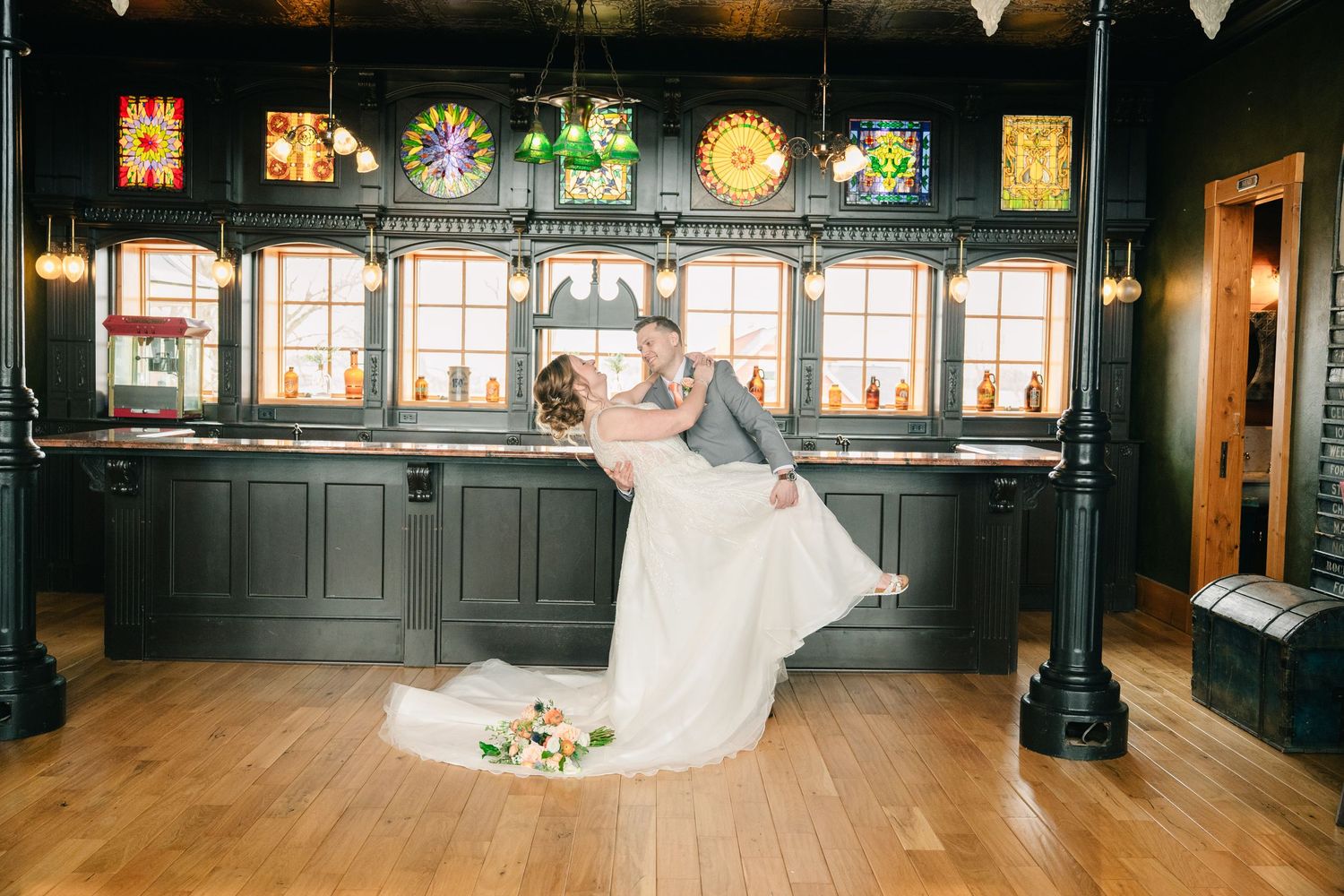 Groom dips bride in elegant wedding venue with stained glass windows and dark wood bar backdrop on hardwood floors.
