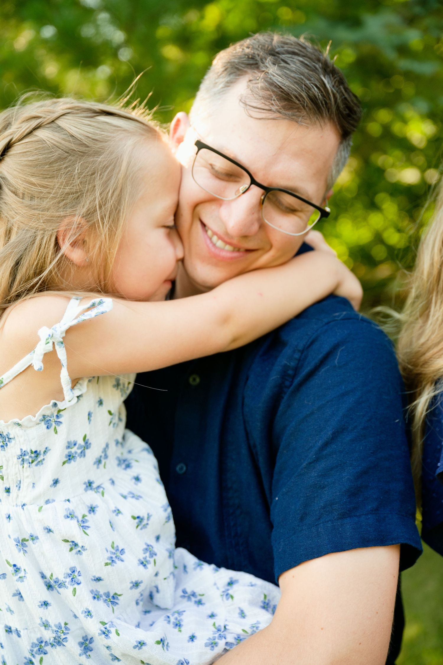 A smiling man with glasses holds a young blonde girl in a floral dress who hugs and kisses his cheek outdoors.