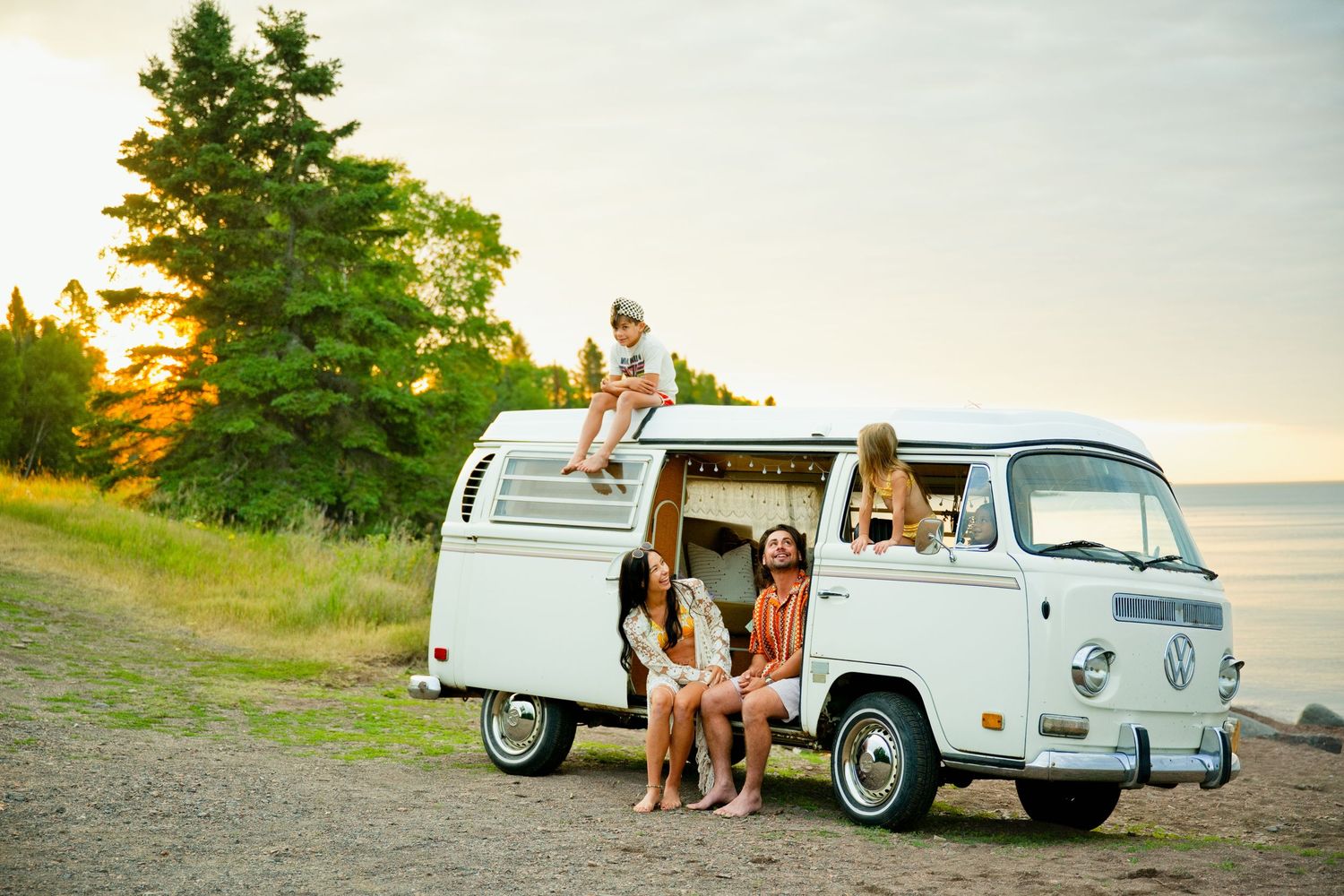 Friends enjoying a road trip in a vintage white VW camper van parked by a scenic lakeside at golden sunset.