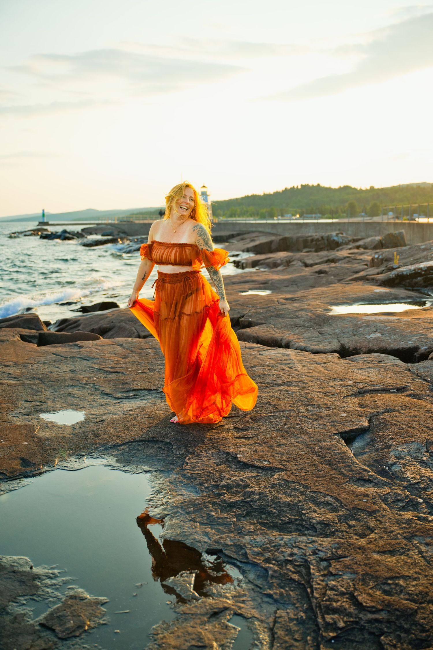 Woman in flowing orange dress stands on rocky shore at sunset with ocean waves behind her.