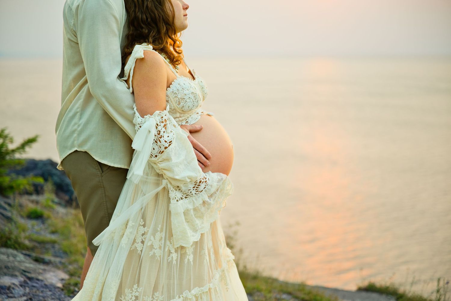 Pregnant woman in lace dress embraced by partner at sunset by the water during maternity photo shoot.