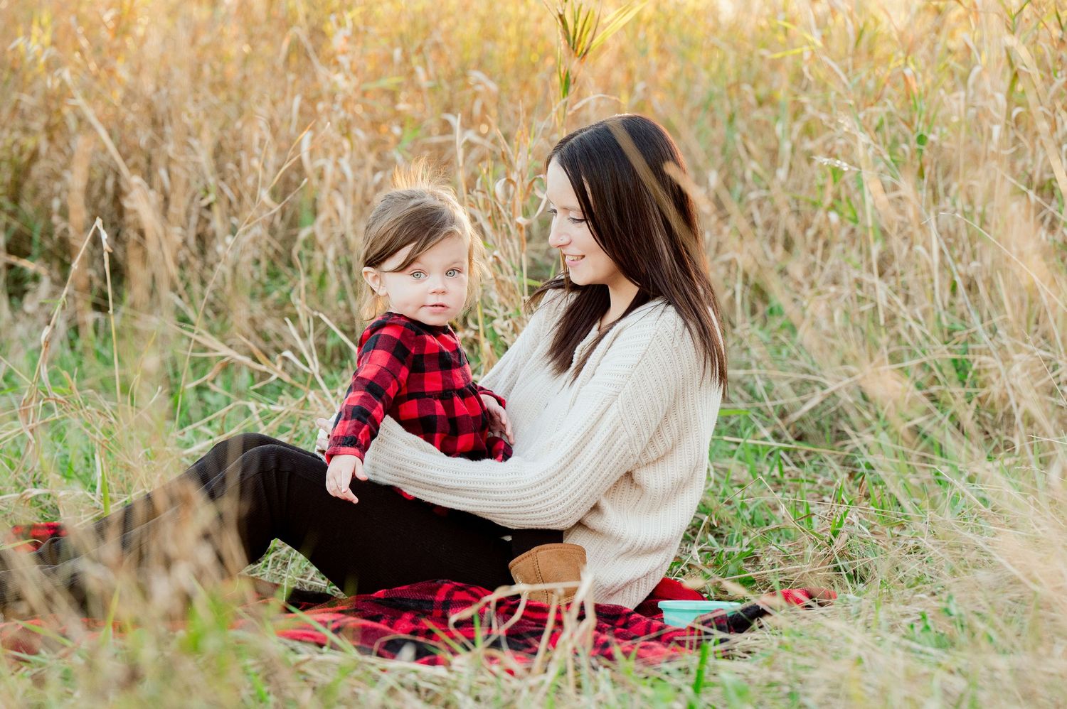 Two people sit together on a red blanket in tall autumn grass wearing cozy sweaters on a fall day.
