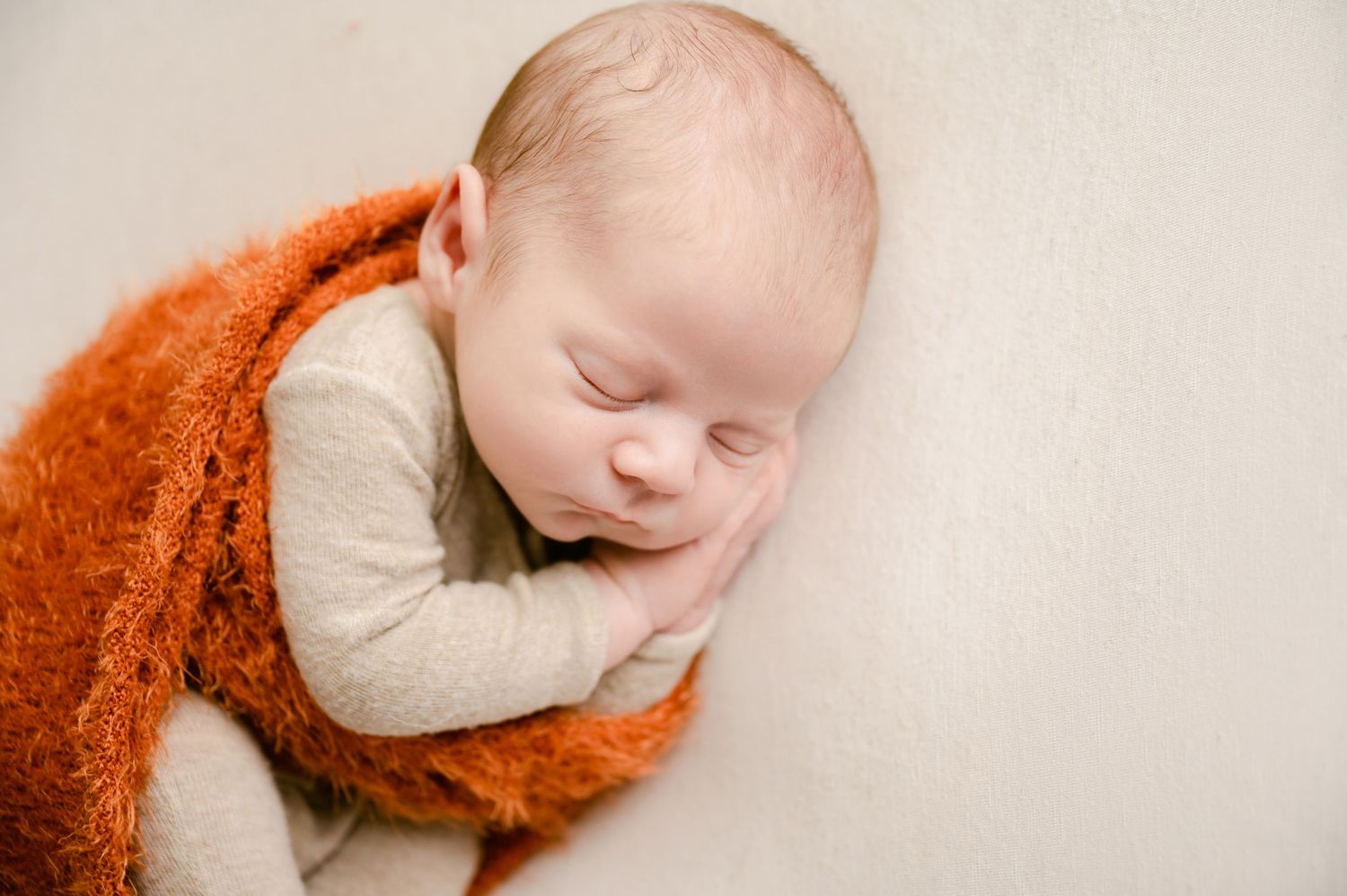Sleeping baby swaddled in soft burnt orange blanket resting peacefully against cream colored backdrop.