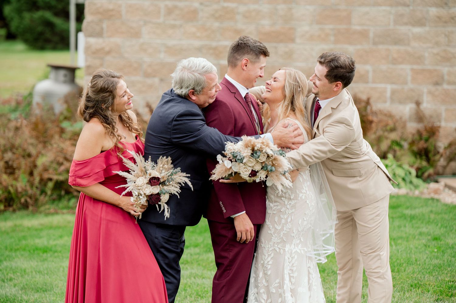A wedding party in burgundy dresses and navy suits poses together on a sunny day by stone walls.