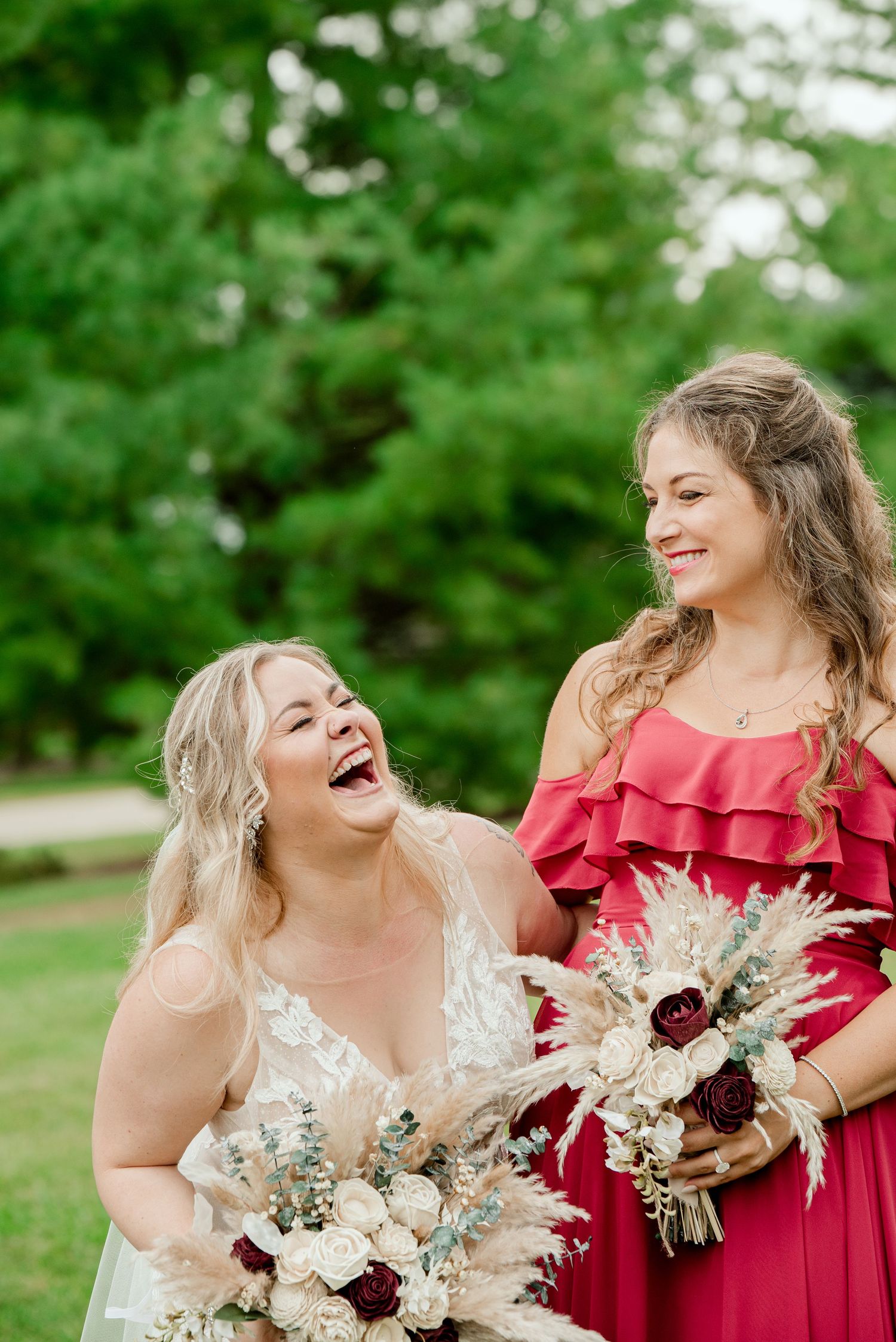 Wedding party members share laughter while holding matching white and burgundy bouquets against green foliage.