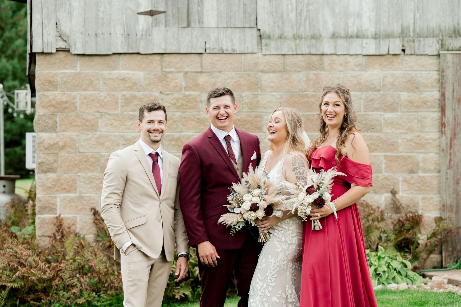 Wedding party poses against rustic barn wall with bride and groom in burgundy attire.