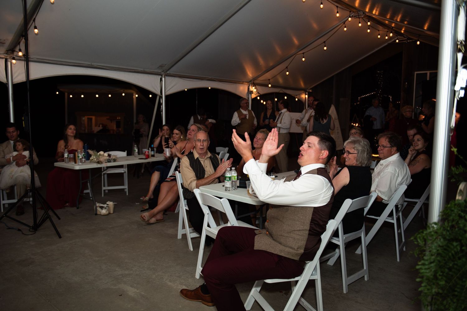 Wedding guests dance and celebrate under a white tent with twinkling lights.