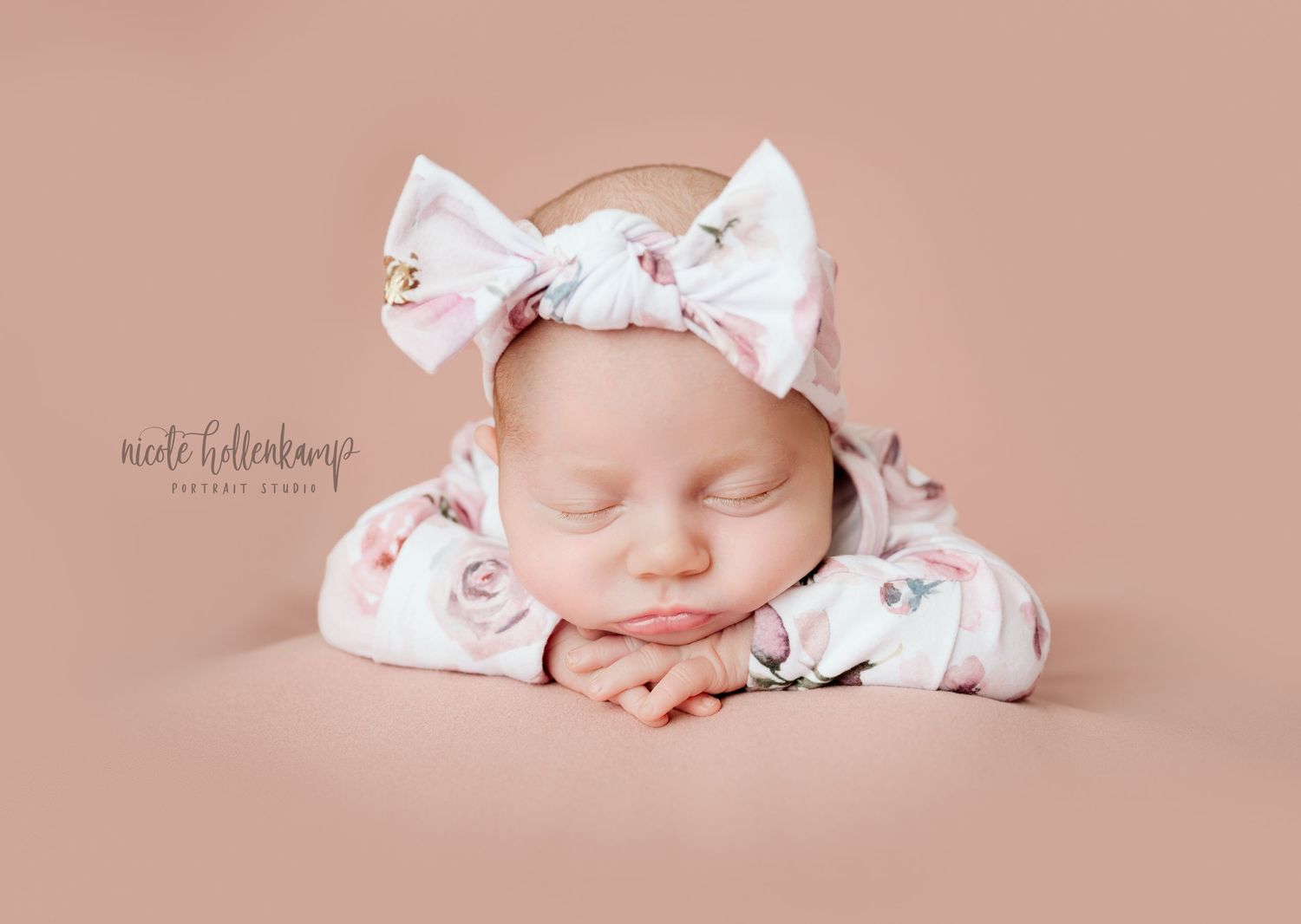 Sleeping newborn wearing floral bow headband on pink background in studio portrait.