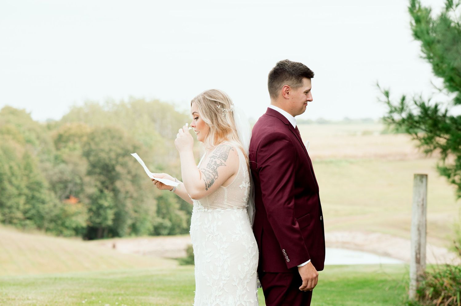 Bride in a lace wedding gown reads a letter while standing back to back with a groom in a burgundy suit in a scenic countryside field.