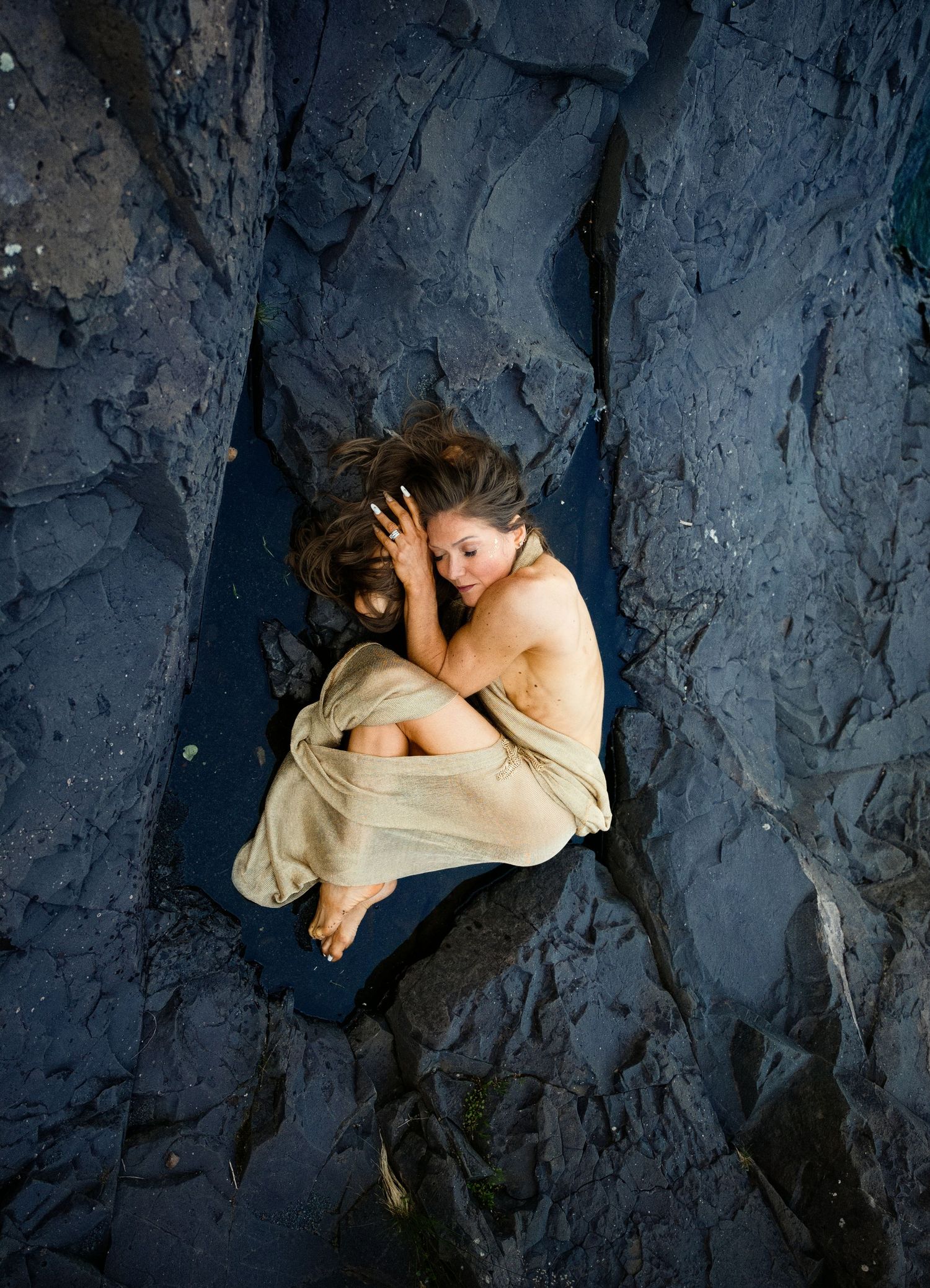 Woman in a draped linen wrap curls up on dark blue volcanic rock, photographed from above in an artistic nature portrait.