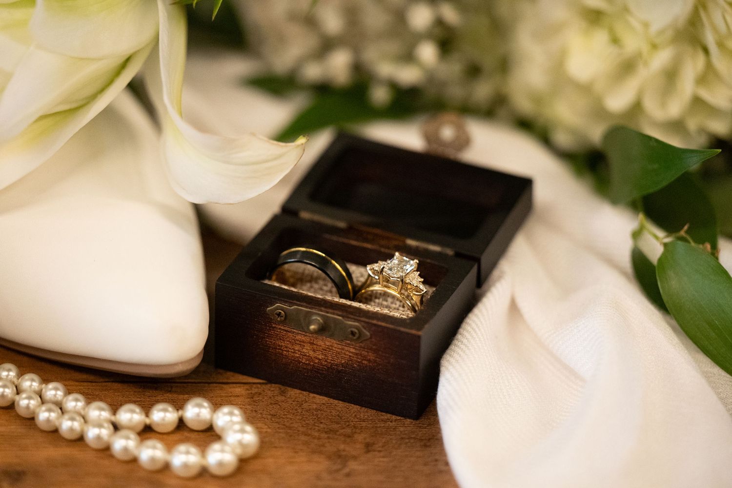 A string of white pearl necklace and wedding rings displayed on a rustic wooden surface with greenery accents at Heart and Soul Chapel Prin.