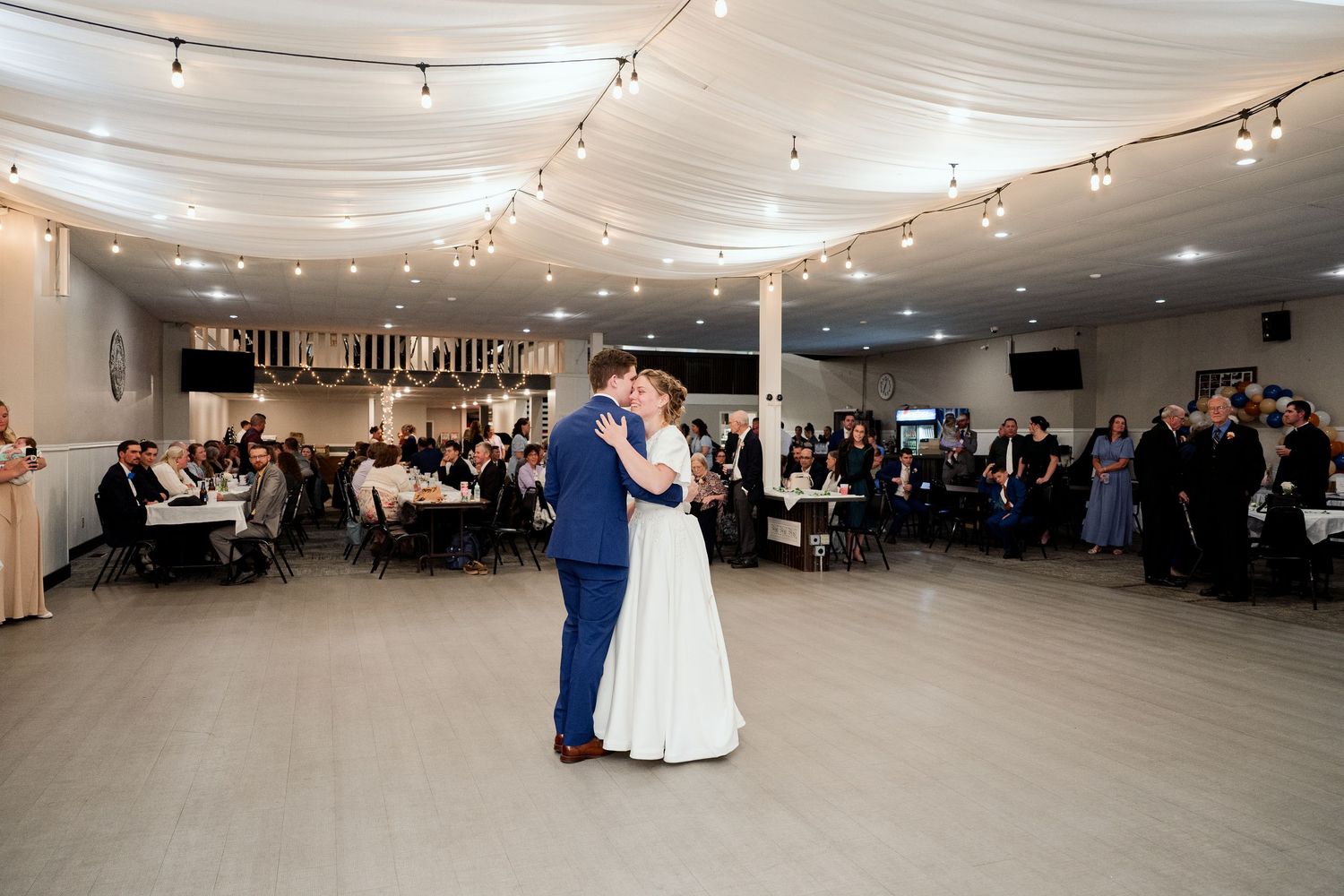 A couple shares their first dance under string lights at an indoor wedding reception venue.