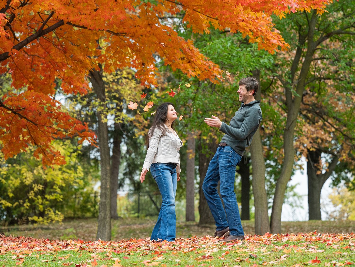 Two figures stand together under bright orange fall foliage in a leaf-covered park landscape at Lake Como. Engagement Sessions Minnesota.